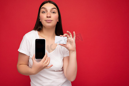 自信、迷人、快乐的黑发年轻女性，穿着休闲白色 T 恤，与红色背景隔离，空间空旷，手持手机，展示智能手机，屏幕上有空屏幕，用于模型和信用卡，看着相机