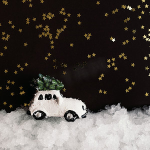 白色闪亮星星摄影照片_一辆微型白色玩具车在雪中的屋顶上挂着一棵圣诞树，背景是黑暗的，上面有闪亮的星星，有复制空间。