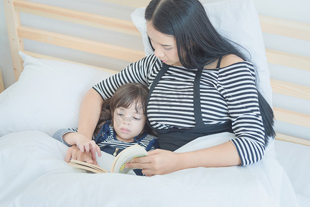 睡前故事摄影照片_幸福的家庭，漂亮的年轻母亲在卧室里给女儿读睡前故事书