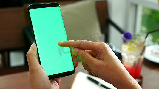 女性手持带空白绿屏的智能手机，在咖啡馆店的绿屏上使用手指滑动，背景为饮料