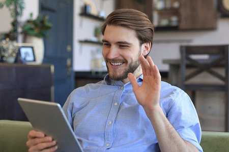 微笑的帅哥在家里拿着和使用带有社交网络的平板电脑，快乐的自由职业者在网上购物，坐在客厅舒适的沙发上