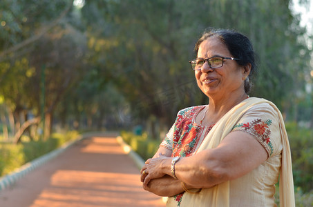 印度新德里摄影照片_夏季，印度新德里，聪明而自信的北印度高级女性穿着灰白色萨尔瓦·卡米兹旁遮普套装，双手交叉/折叠在公园里站着、思考、看着别处