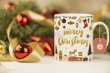 圣诞杯的特写，上面挂着空茶标签，周围有松枝、红色小饰品和木制背景上的缎带金丝带，具有散景效果