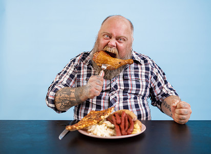 超重的风趣男人在浅蓝色背景的餐桌上吃熏鸡腿