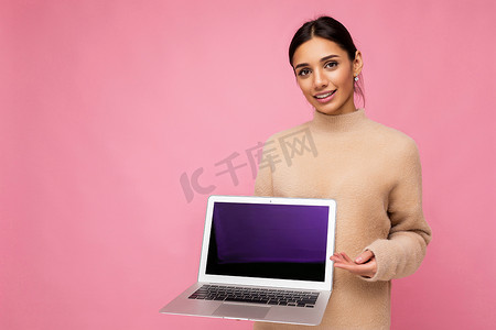 粉色墙壁摄影照片_美丽的年轻女子，深色头发，看着相机，手持电脑笔记本电脑，显示屏空空如也，带有模拟和复制空间，穿着浅色毛衣，与粉色墙壁背景隔离