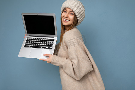 特写侧面照片拍摄美丽迷人迷人漂亮微笑快乐的年轻深色金发女人在冬天温暖的针织米色帽子拿着电脑笔记本电脑看着相机穿着米色冬季毛衣隔离在浅蓝色墙壁背景
