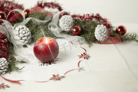 圣诞节设置：前景上有交叉屏幕效果的红色点燃蜡烛，周围环绕着松树枝、红色小饰品、红色和白色丝带、圣诞桌布上的白色松果，具有散景效果