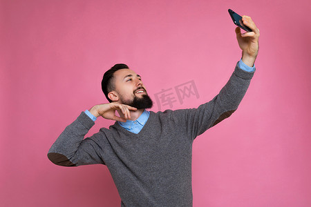 正面英俊的年轻黑发未剃须男子的照片，留着胡子，穿着休闲的灰色毛衣和蓝色衬衫，隔离在粉红色背景墙上，手持智能手机自拍照片，看着手机屏幕显示