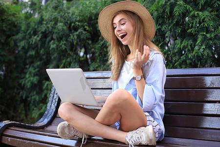 年轻漂亮的女人穿着夏装，坐在城市街道的长椅上，在笔记本电脑上工作。