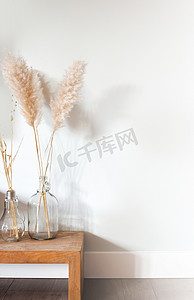 装饰草摄影照片_靠近白色背景的木桌上玻璃花瓶中的干潘帕斯草，现代明亮的家居室内装饰，复制空间