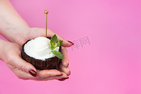 白色冰淇淋球摄影照片_香草冰淇淋球放在新鲜椰子的一半里，手里拿着薄荷叶装饰，粉色背景上放着勺子