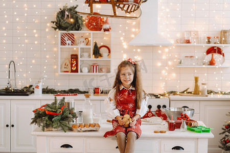 圣诞桌面摄影照片_圣诞厨房里一个快乐的小女孩坐在桌子上，手里拿着饼干
