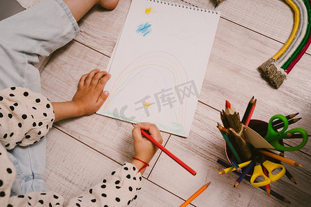 儿童小彩虹摄影照片_一个小女孩坐在地板上，用彩色铅笔在速写本上画画。