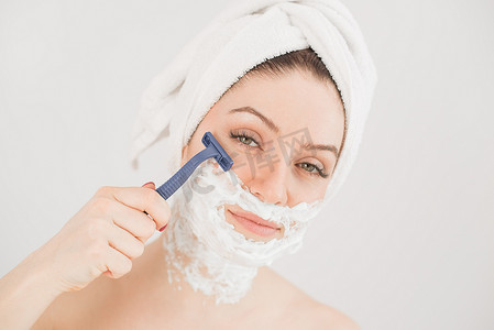 脸上的泡沫摄影照片_头戴毛巾、脸上剃须泡沫的快乐白人女性拿着白色背景上的剃刀