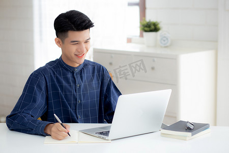 工作中计划摄影照片_年轻的亚洲商人在笔记本上写作，以计划在家里的办公桌上工作和使用笔记本电脑，记下金融、男性学习和学习、商业和沟通概念。