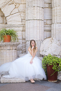 背景墙柱子摄影照片_白种人迷人的新娘坐在古老的柱子附近，穿着白色的连衣裙。