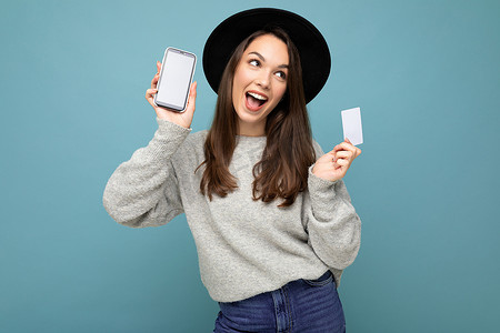 蓝色背景中，身穿黑色帽子和灰色毛衣的迷人、快乐的黑发年轻女性手持信用卡和手机的照片，显示空白，供模特看向侧面
