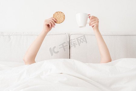 捧着杯子摄影照片_女性双手捧着一杯咖啡和饼干躺在床上的毯子上方。