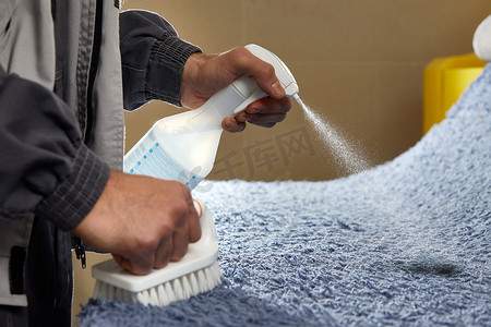 脏的污渍摄影照片_男子在灰色地毯上喷洒洗涤剂以去除专业清洁服务中的污渍