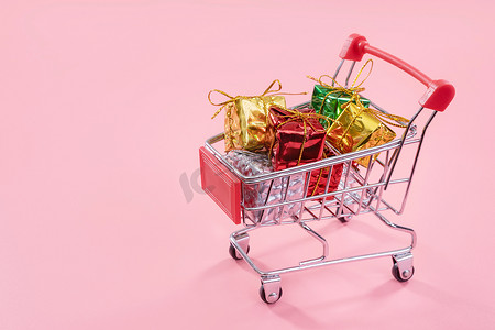 年度销售摄影照片_年度销售、圣诞购物季概念 — 迷你红色购物车手推车，里面装满了淡粉色背景中突显的礼盒，复制空间，特写