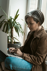 中年有魅力的女商人坐在免费工作区或工作站的窗台上，从事笔记本电脑的工作。