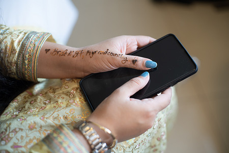 年轻的印度女孩穿着白金礼服，准备举行婚礼，手里拿着手机，上面用指甲花纹身写着“新娘是最好的朋友”