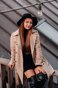 一位穿着米色外套和黑色帽子的时尚年轻女子坐在市中心的屋顶上。