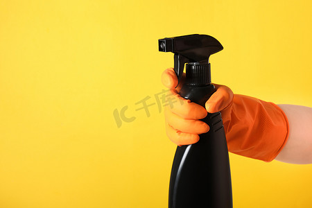 橙色手套里的一只手拿着黄色背景上的黑色清洁液喷雾瓶