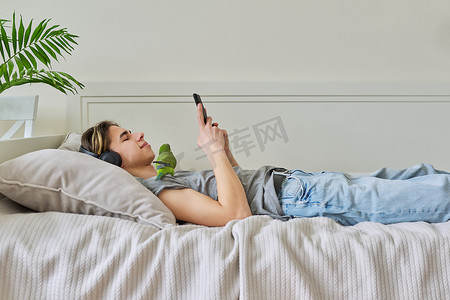 男性青少年与鹦鹉躺在家里的床上，戴着耳机和智能手机