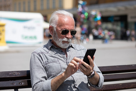 老人技术摄影照片_老人在智能手机上使用互联网应用程序，坐在长凳上