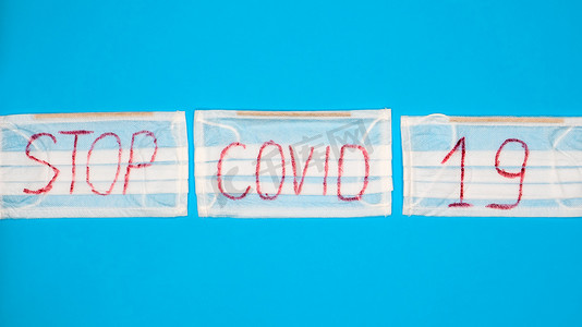 英文字母字母摄影照片_典型的 3 层白色外科口罩，带有橡胶耳钩，用蓝色背景上的英文大写字母 Stop Covid-19 盖住嘴巴和鼻子。