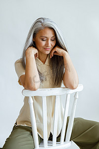 灰白的头发摄影照片_浅色背景下，头发灰白的中年亚洲女性向后坐在椅子上