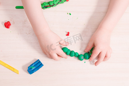 一个小孩在木桌上用绿色橡皮泥雕刻毛毛虫，发展手的精细运动技能，在家里和孩子一起玩，前景用橡皮泥毛毛虫
