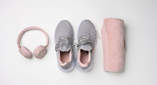 一双灰色纺织运动鞋、无线耳机和一条白色背景的粉色纺织毛巾。