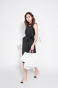 黑色购物摄影照片_美丽的亚洲女人穿着黑色连衣裙，提着购物袋，站在白色的上方。