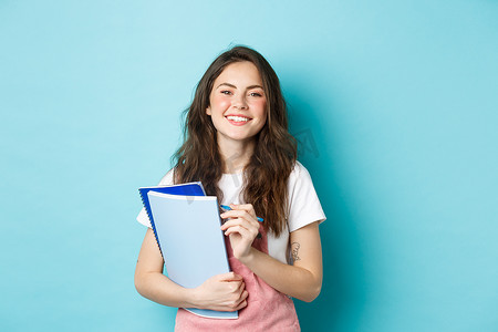 快乐的年轻女学生拿着课程笔记本，对着镜头微笑，穿着春装站在蓝色背景下