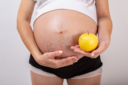 孕妇触摸她的大肚子，手里拿着白色背景的黄色苹果的图像。