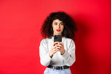 卷发的年轻女性，在智能手机上录制视频，在手机上拍照，看着相机，站在红色背景上