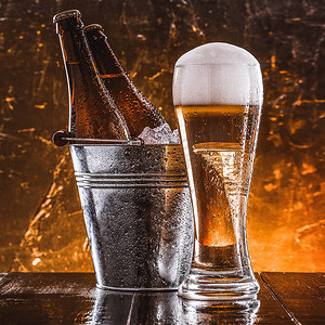两瓶啤酒装在一个桶里，里面装着冰，还有一杯啤酒，泡沫丰富，紧挨着深色背景