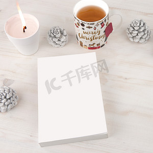 从顶部看书的圣诞背景：空白封面的书，上面点着白蜡烛，圣诞装饰的茶杯，浅色木桌上放着白松果
