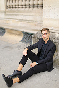 年轻的欧洲男子坐在人行道上，身穿黑色西装。