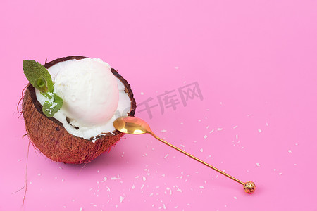 白色冰淇淋球摄影照片_新鲜椰子中的香草冰淇淋球，一半用薄荷叶装饰，粉红色背景上有勺子