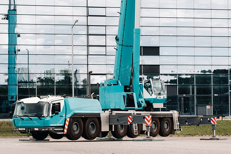 起重机维修摄影照片_大型蓝色卡车起重机随时准备在大型现代建筑旁边的平台上操作液压支架。
