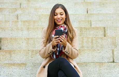 微笑年轻漂亮的女人使用智能手机坐在公共公园的楼梯上。