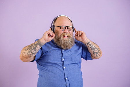 留着胡子、超重的快乐男人戴着紫色背景的耳机听音乐