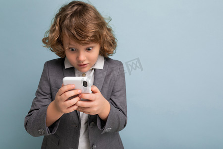 短卷发男孩摄影照片_照片中，英俊的卷发男孩穿着灰色西装，手持并使用蓝色背景中隔离的手机，看着智能手机通信 vie 短信