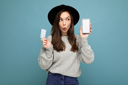 照片中，迷人的黑发年轻女性身穿黑色帽子和灰色毛衣，与蓝色背景隔离，手持信用卡和手机，显示屏空空如也，供模特看向侧面