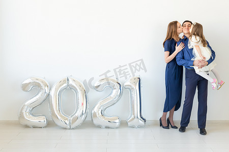 年轻幸福的家庭母亲、父亲和女儿站在白色背景上形状像数字 2021 的气球旁边。