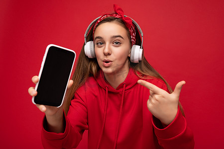 身穿红色连帽衫、红色背景中突显的迷人、快乐、惊奇的黑发女孩手持并展示智能手机，显示屏空空如也，戴着白色无线耳机，听着有趣的音乐，看着相机，指着设备