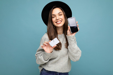 身穿黑色帽子和灰色毛衣的美丽正面微笑的黑发年轻女性，在蓝色背景中被隔离，手持信用卡和手机，显示屏空空如也，用于模拟看着相机